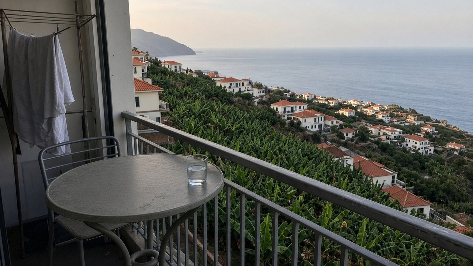 A closed laptop and a glass of water on a balcony table above banana terraces and the sea