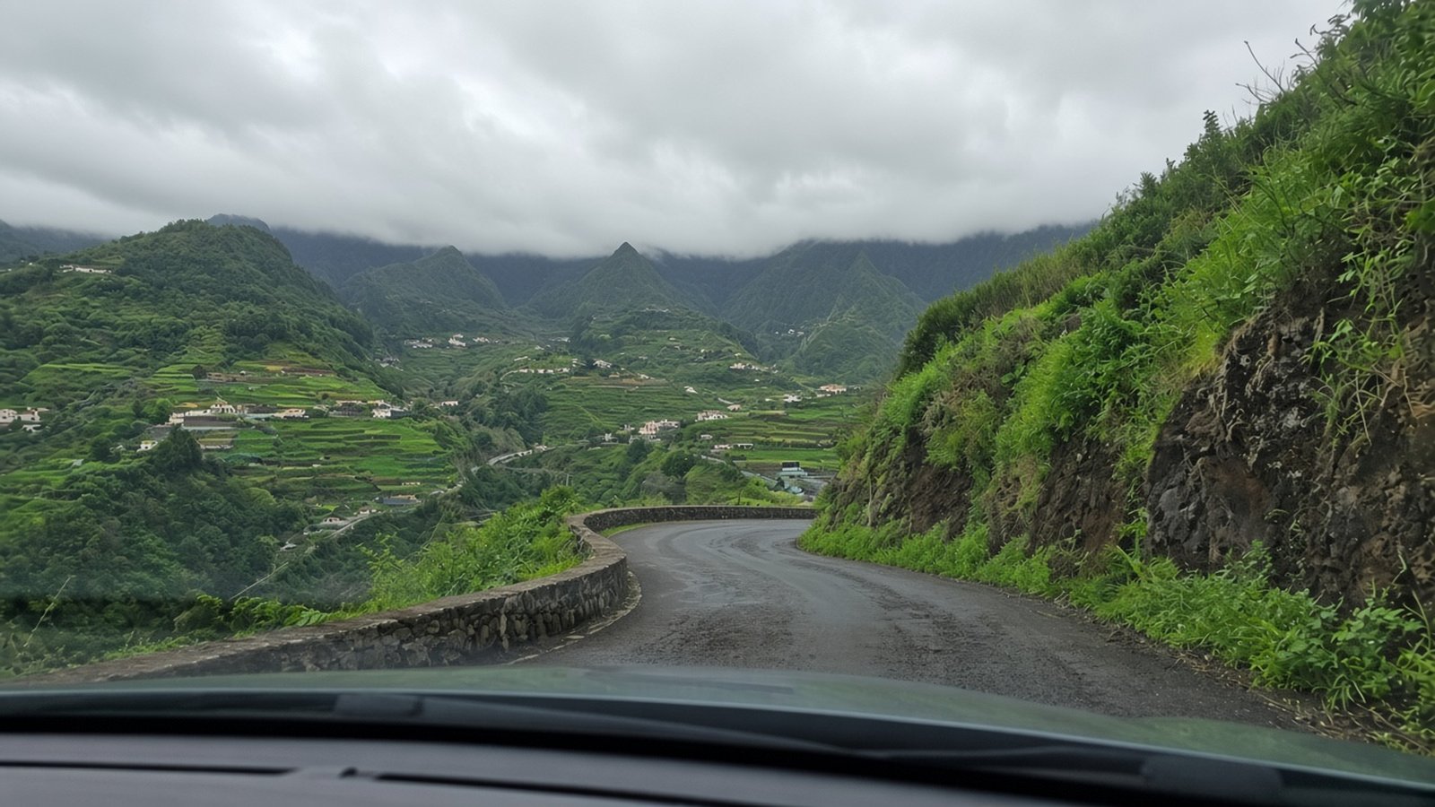 View through a car windscreen of a narrow mountain road curving past terraced green valleys under low cloud