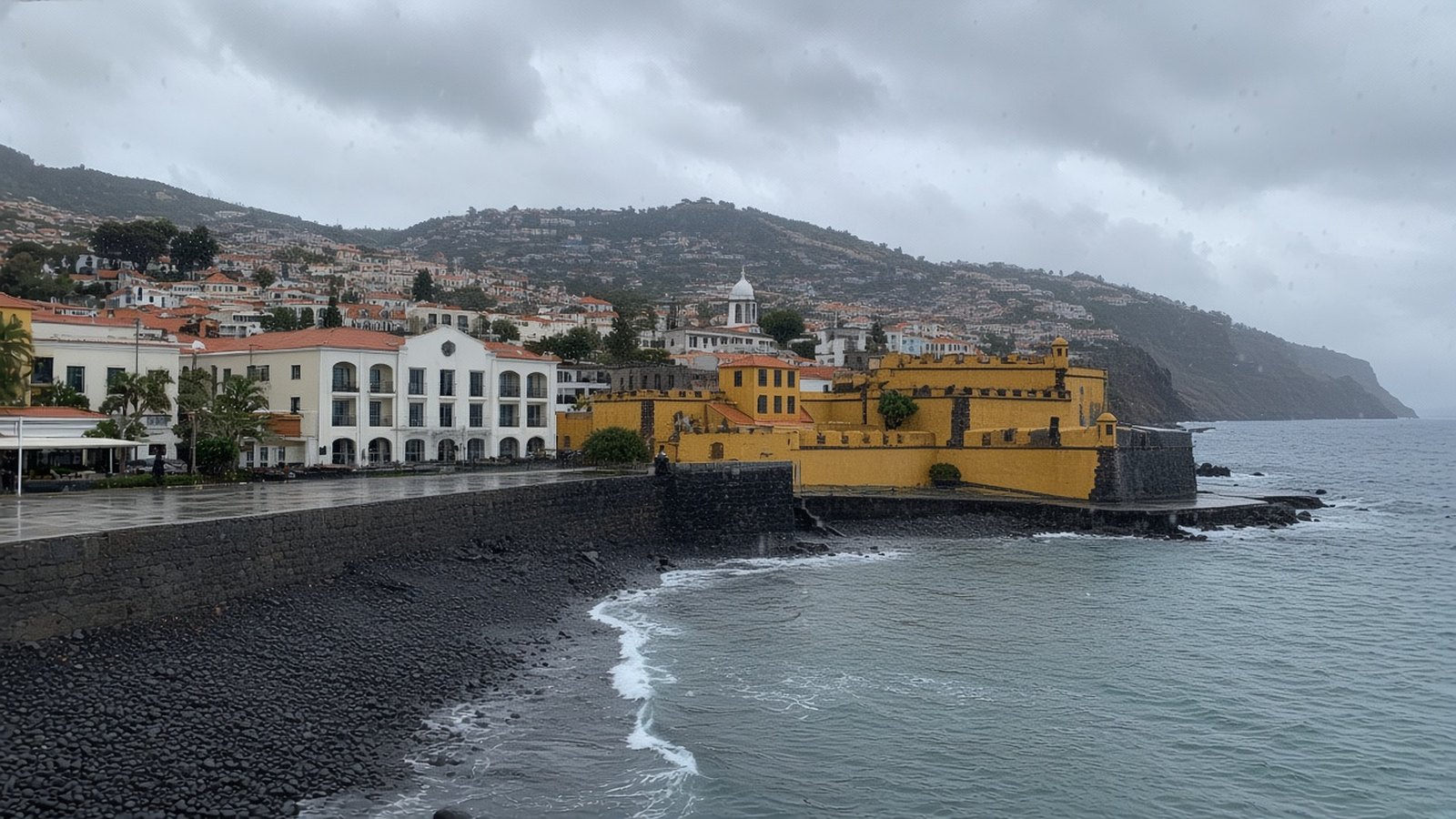 Rain on the lens over Funchal's ochre São Tiago fort, the white hotel beside it, and wet basalt at the Zona Velha shore