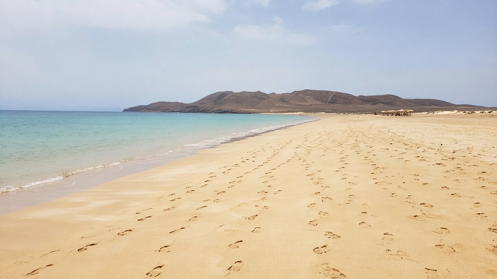 Footprints on a long golden sand beach beside turquoise water, with brown hills on the horizon